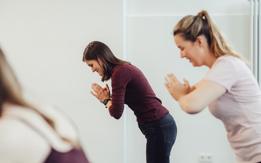 Zwei Frauen stehen nebeneinander in einer Yogaposition und halten ihre Hände vor sich zusammen