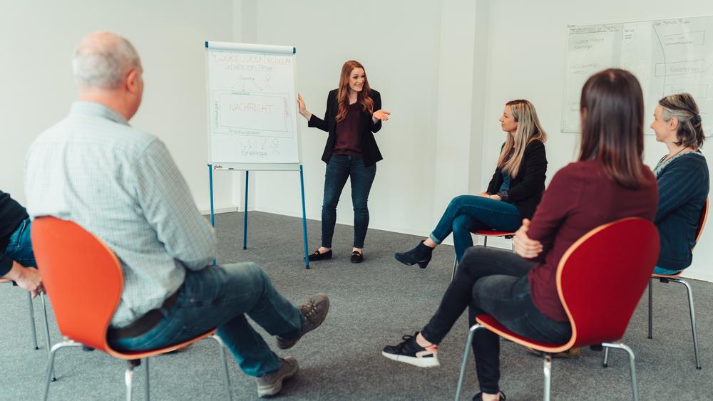 Gruppentherapie mit mehrern Personen, die im Kreis sitzen und einer Frau neben einem Plakat