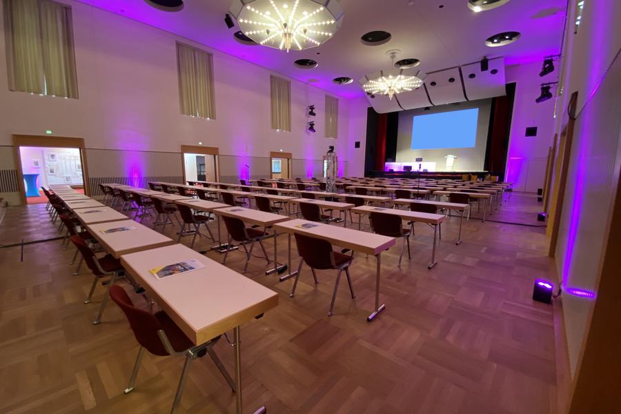 Großer beleuteter Saal mit Tischreihen und einer großen Leinwand