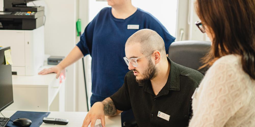 Drei Personen arbeiten gemeinsam an einem Computerarbeitsplatz und besprechen Unterlagen