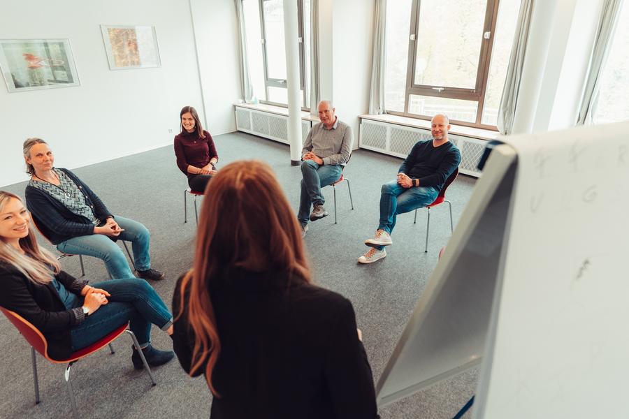 Gruppentherapie mit mehrern Personen, die im Kreis sitzen und einer Frau neben einem Plakat