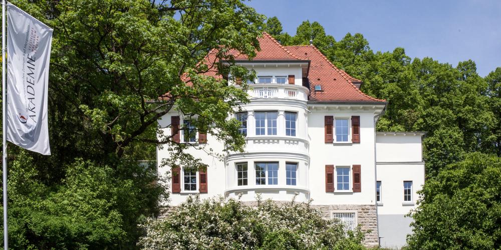 Weißes Akademiegebäude teils abgedeckt von umliegenden Bäumen