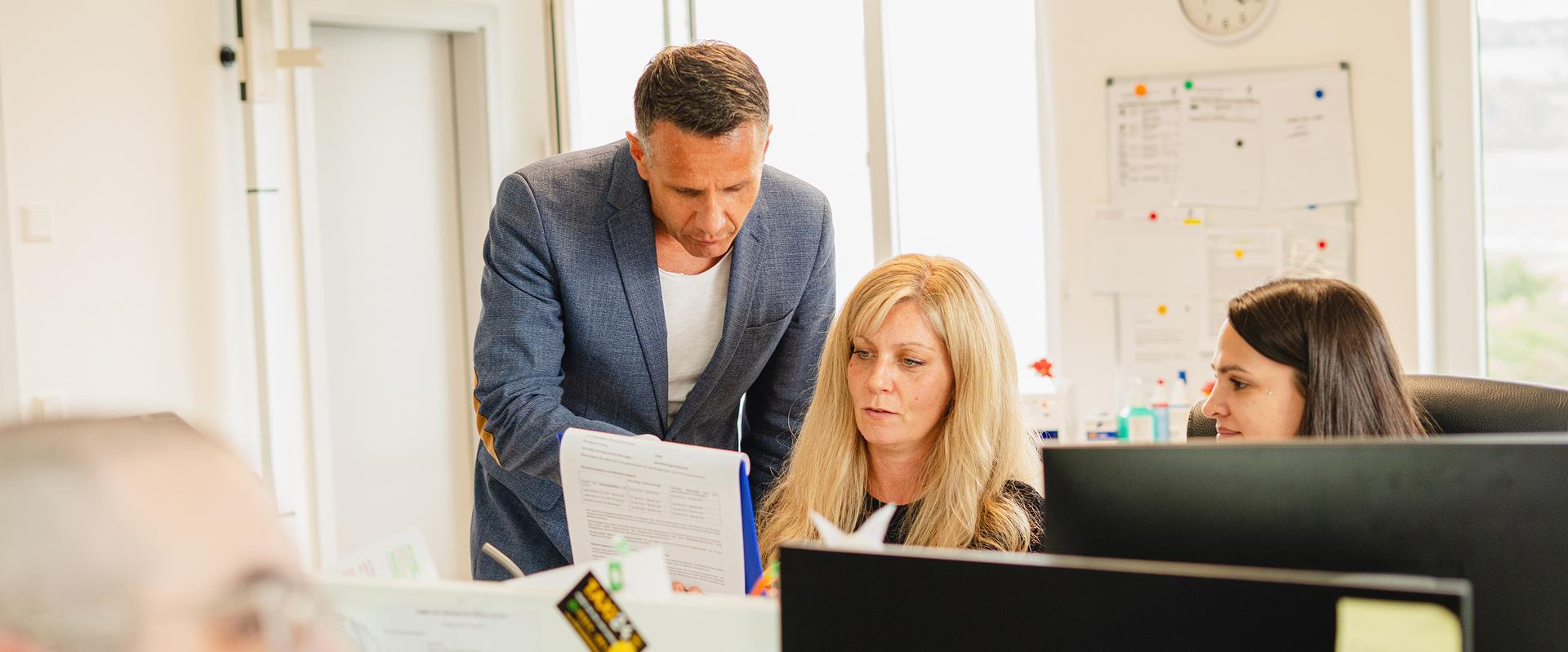 Drei Personen arbeiten gemeinsam an einem Computerarbeitsplatz und besprechen Unterlagen