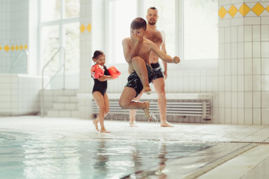 Junge springt ins Schwimmbecken, Mädchen und Vater stehen am Beckenrand