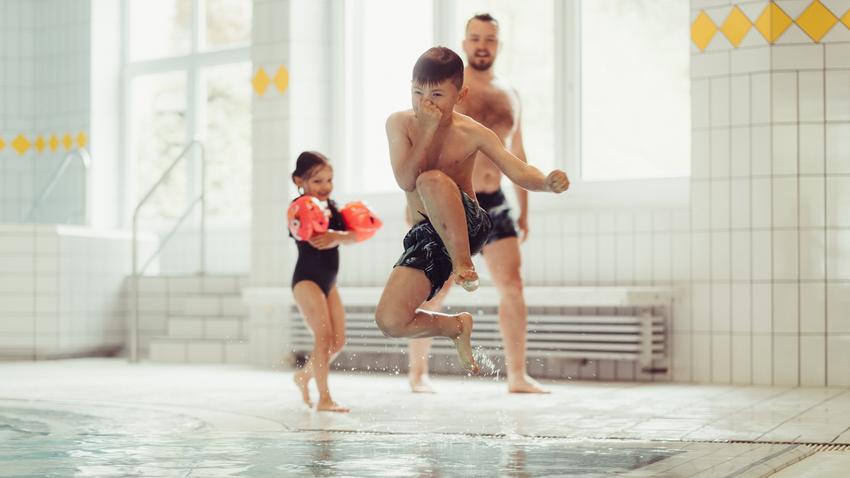 Junge springt ins Schwimmbecken, Mädchen und Vater stehen am Beckenrand